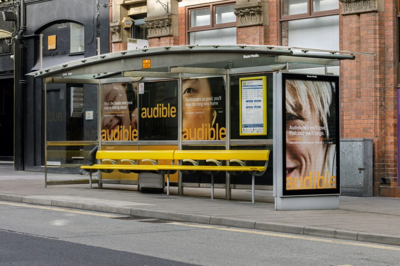 Wide view of an Audible bus shelter wrap installation showing multiple panels and a freestanding poster in a UK streetscape.