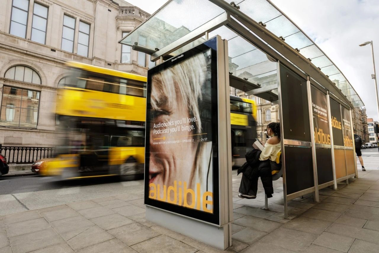 Audible bus shelter poster featuring podcast messaging with a yellow bus passing in the background.