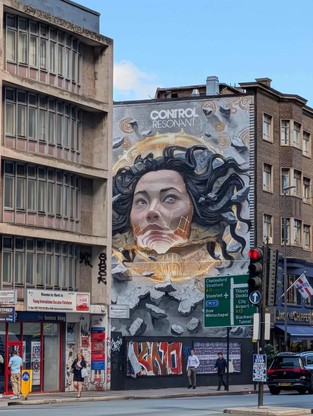 Large Control Resonant mural on a London building showing a woman's face surrounded by fractured debris, dark flowing forms and circular symbols above UK road signs.