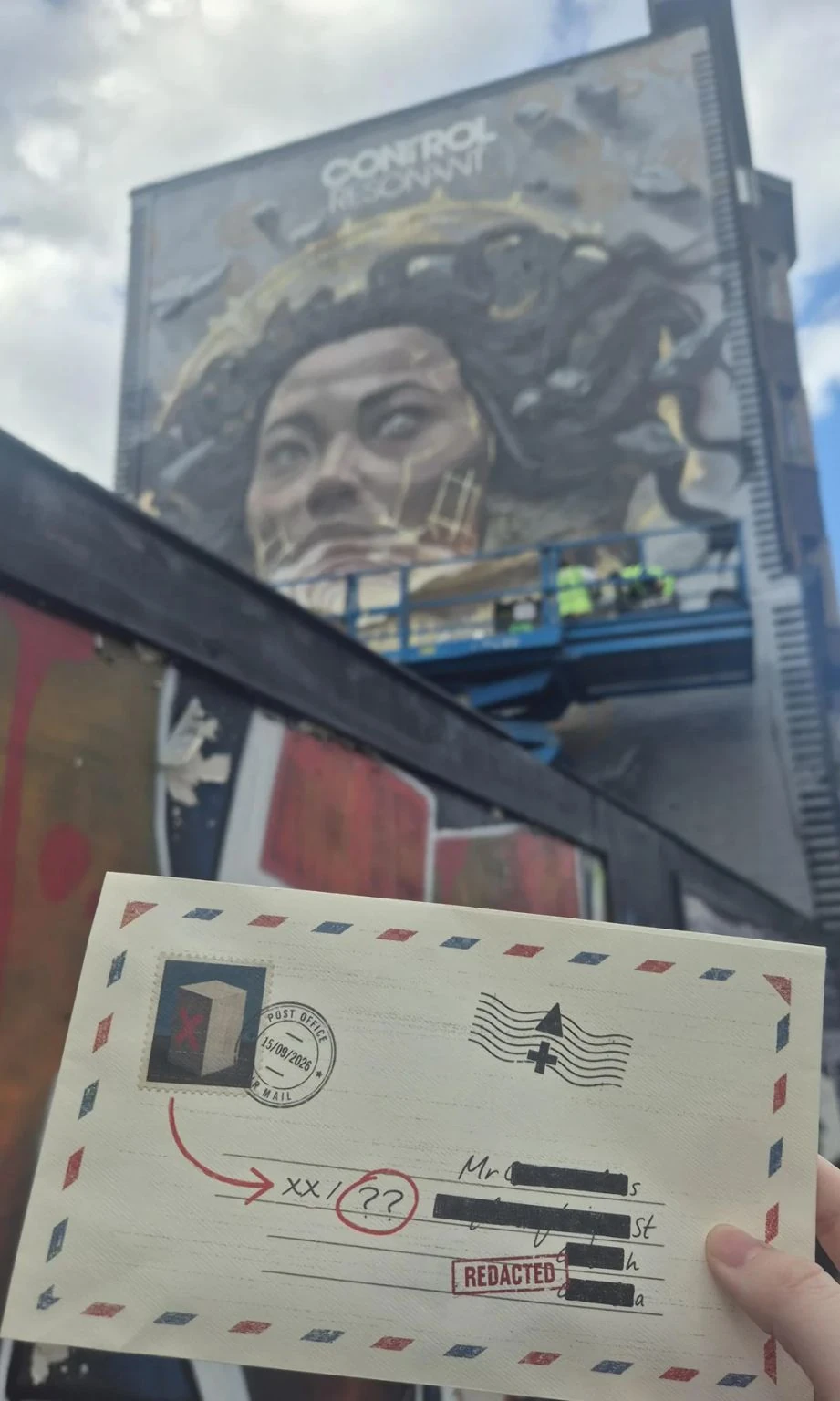 Hand holding a redacted airmail-style envelope dated 15 September 2026 in front of the Control Resonant London mural, with blacked-out address details and a red REDACTED stamp.