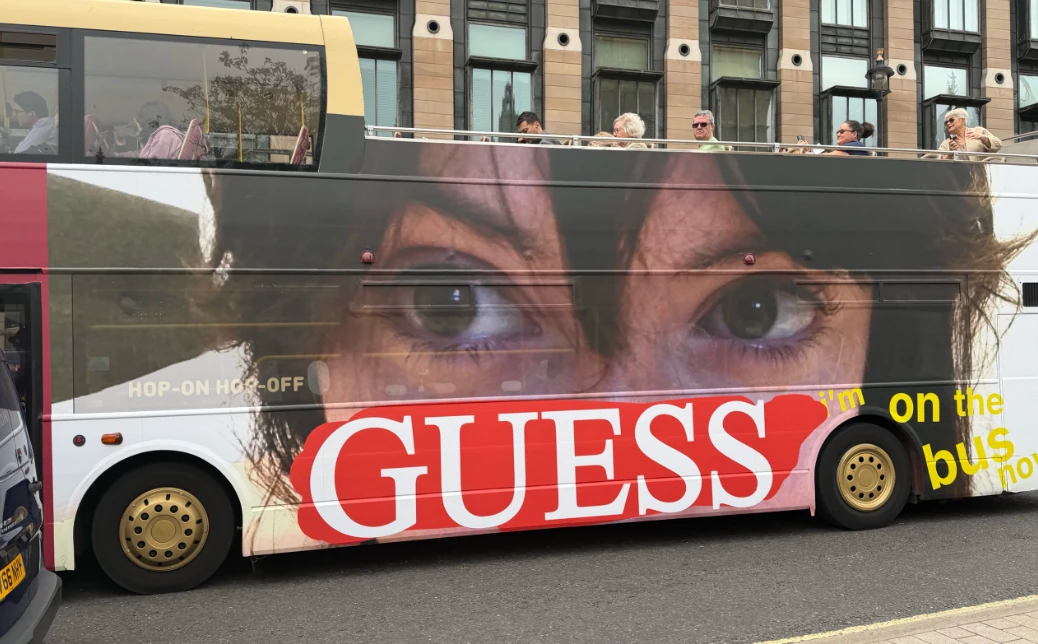 Side view of a Guess Girls campaign double-decker bus in London featuring an oversized portrait, the Guess logo and lo-fi campaign typography.