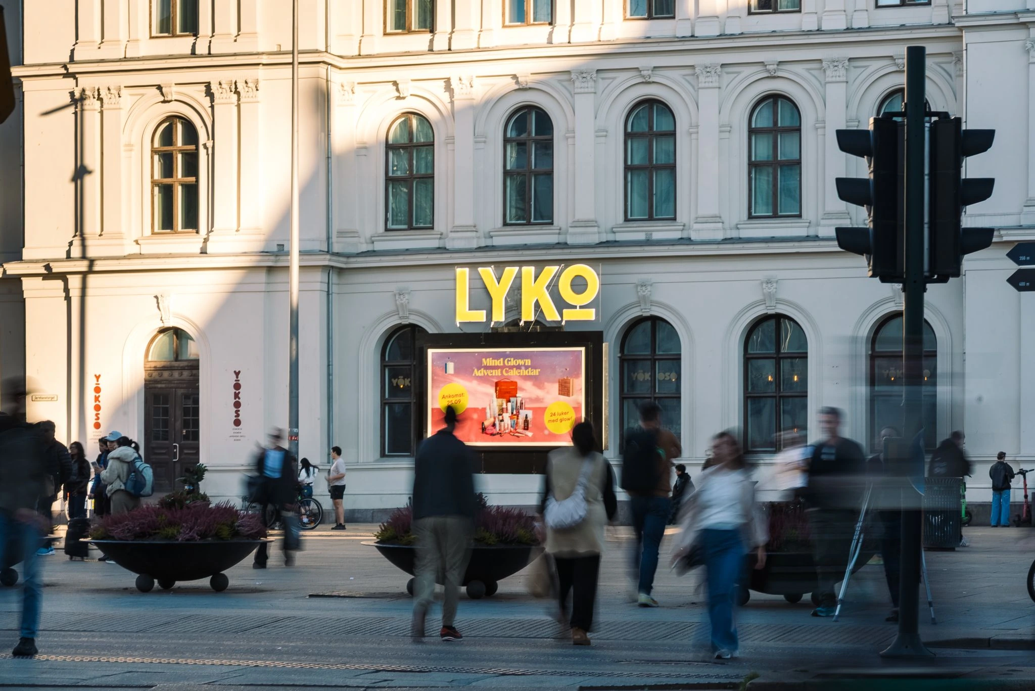 Daytime street scene at Jernbanetorget in Oslo showing LYKO's special build with glowing LYKO branding and a digital screen promoting the Mind Glow Advent Calendar while pedestrians cross the plaza.