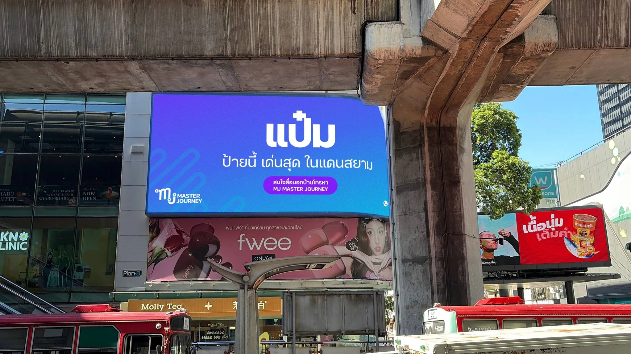 A large blue MJ Master Journey billboard displayed in a busy Bangkok commercial district beneath elevated transit infrastructure, surrounded by street traffic and retail signage.