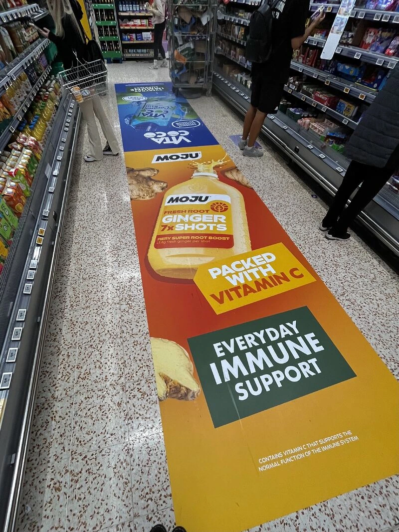 Large MOJU floor graphic inside Morrisons promoting Ginger 7x Shots, vitamin C and everyday immune support.
