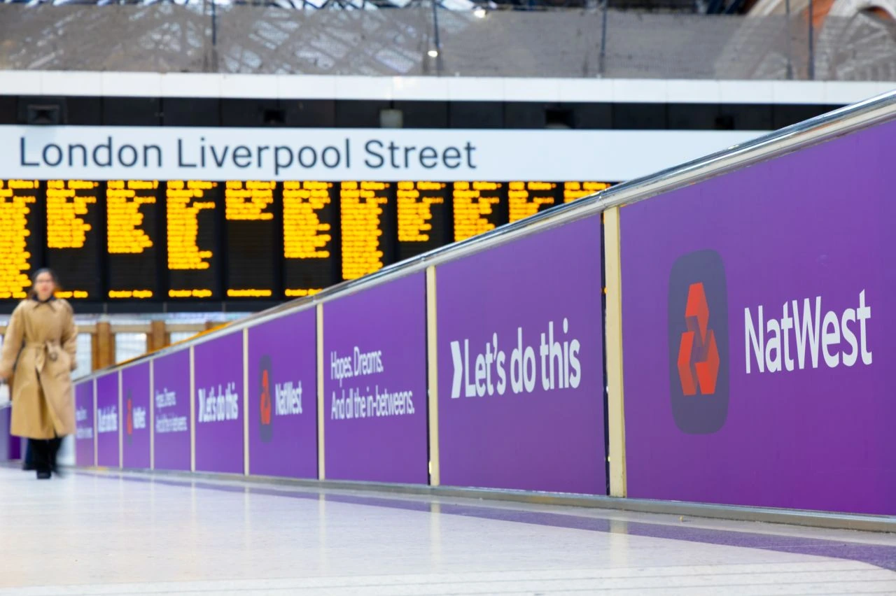 Purple NatWest Let’s do this branding running along a walkway inside London Liverpool Street station beside the main departure boards.