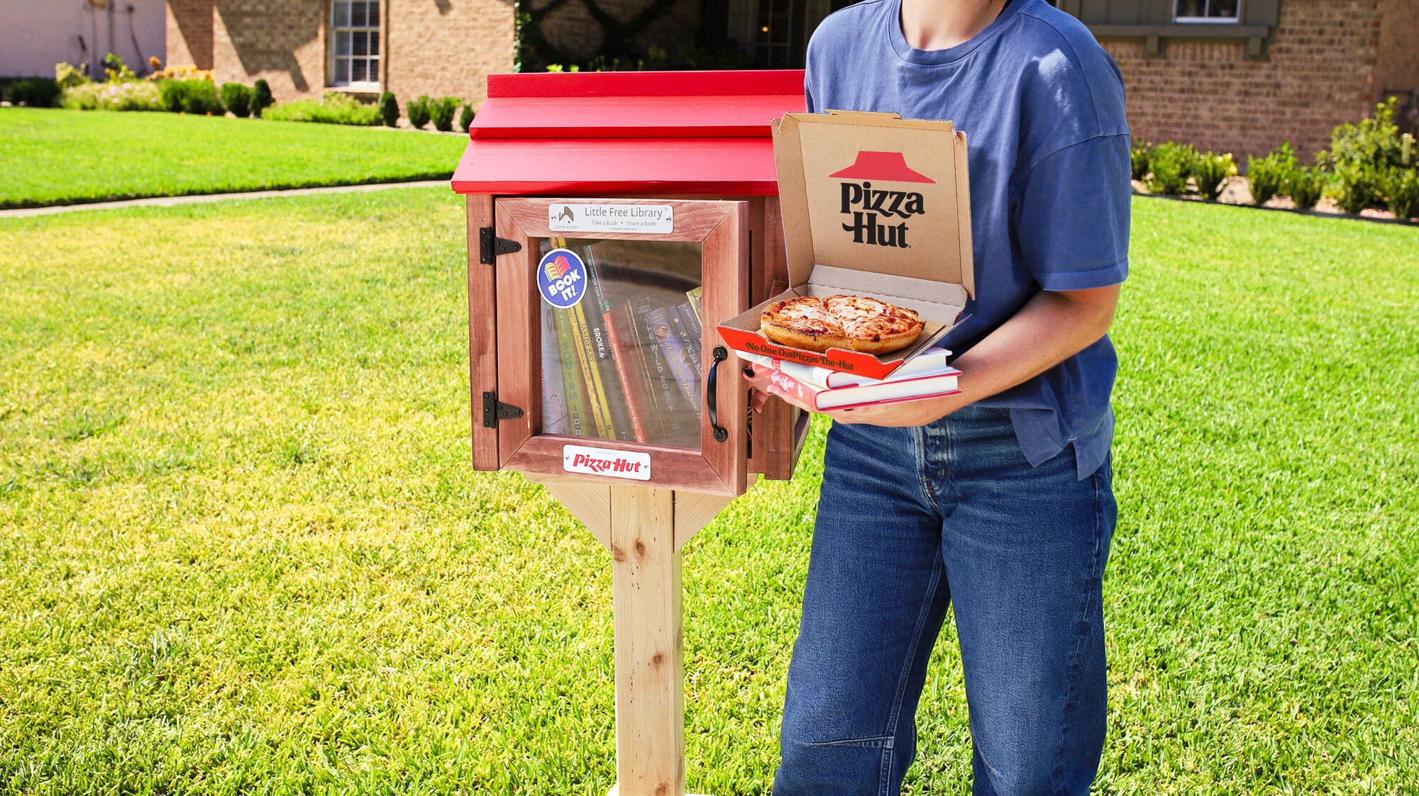 Person holding books and a Pizza Hut Personal Pan Pizza next to a Little Hut Library.