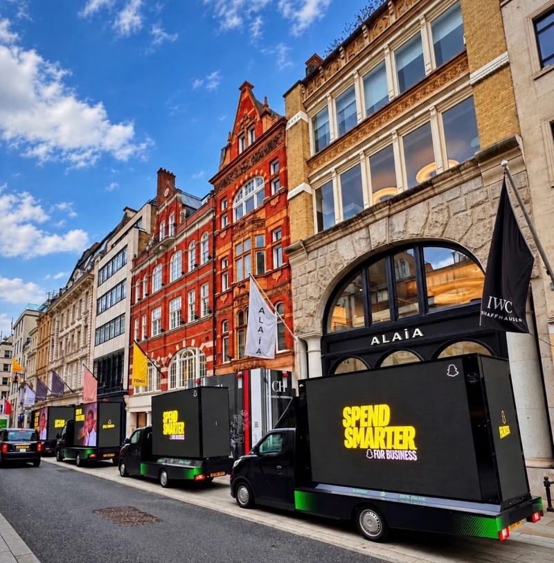 Three Snapchat mobile digital billboard vans parked on a London shopping street near luxury storefronts, showing the yellow Spend Smarter message.