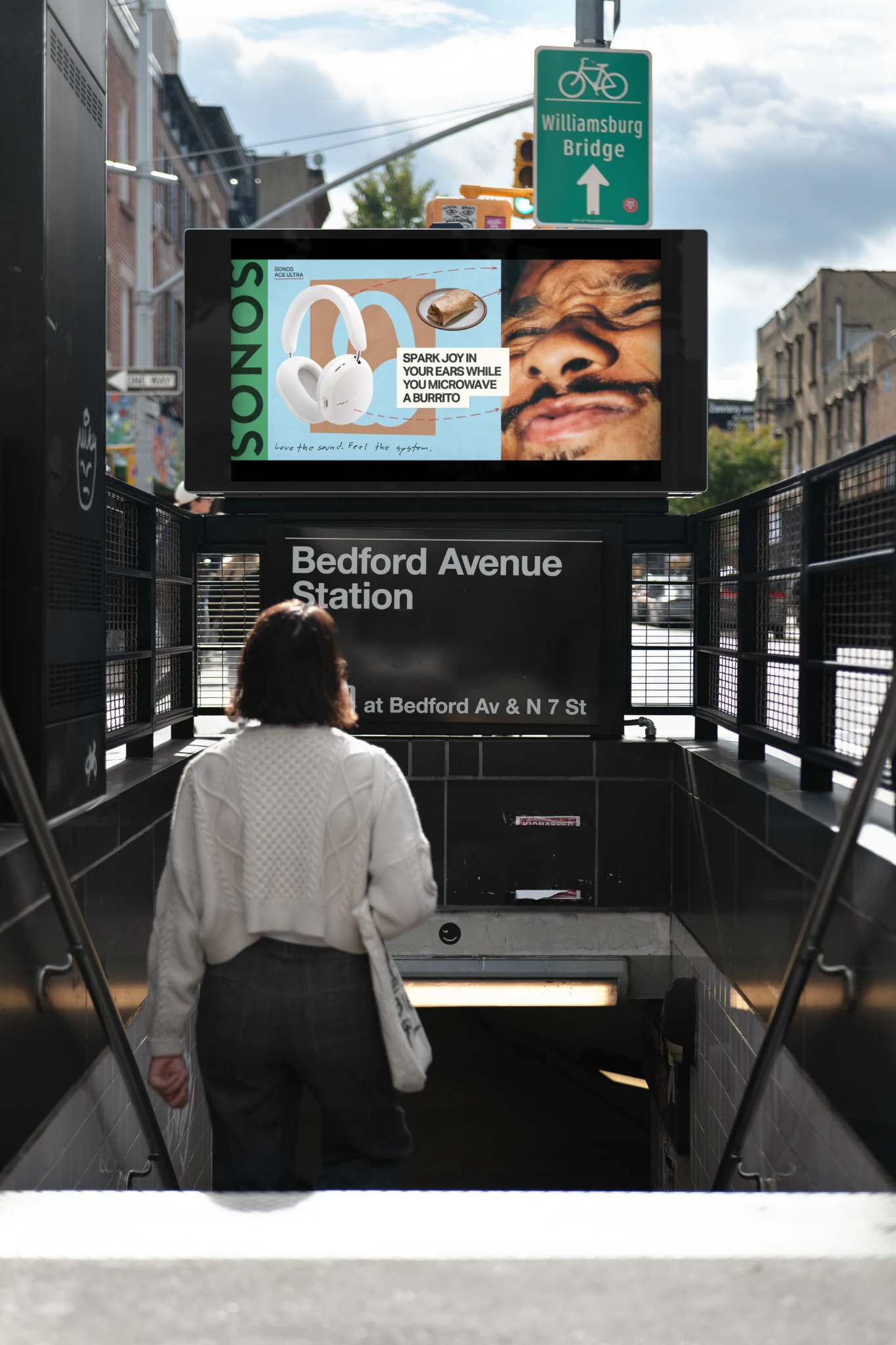 Digital Sonos Ace Ultra advertisement above the Bedford Avenue Station entrance in Williamsburg, Brooklyn, featuring white headphones, a burrito and the headline Spark joy in your ears while you microwave a burrito.