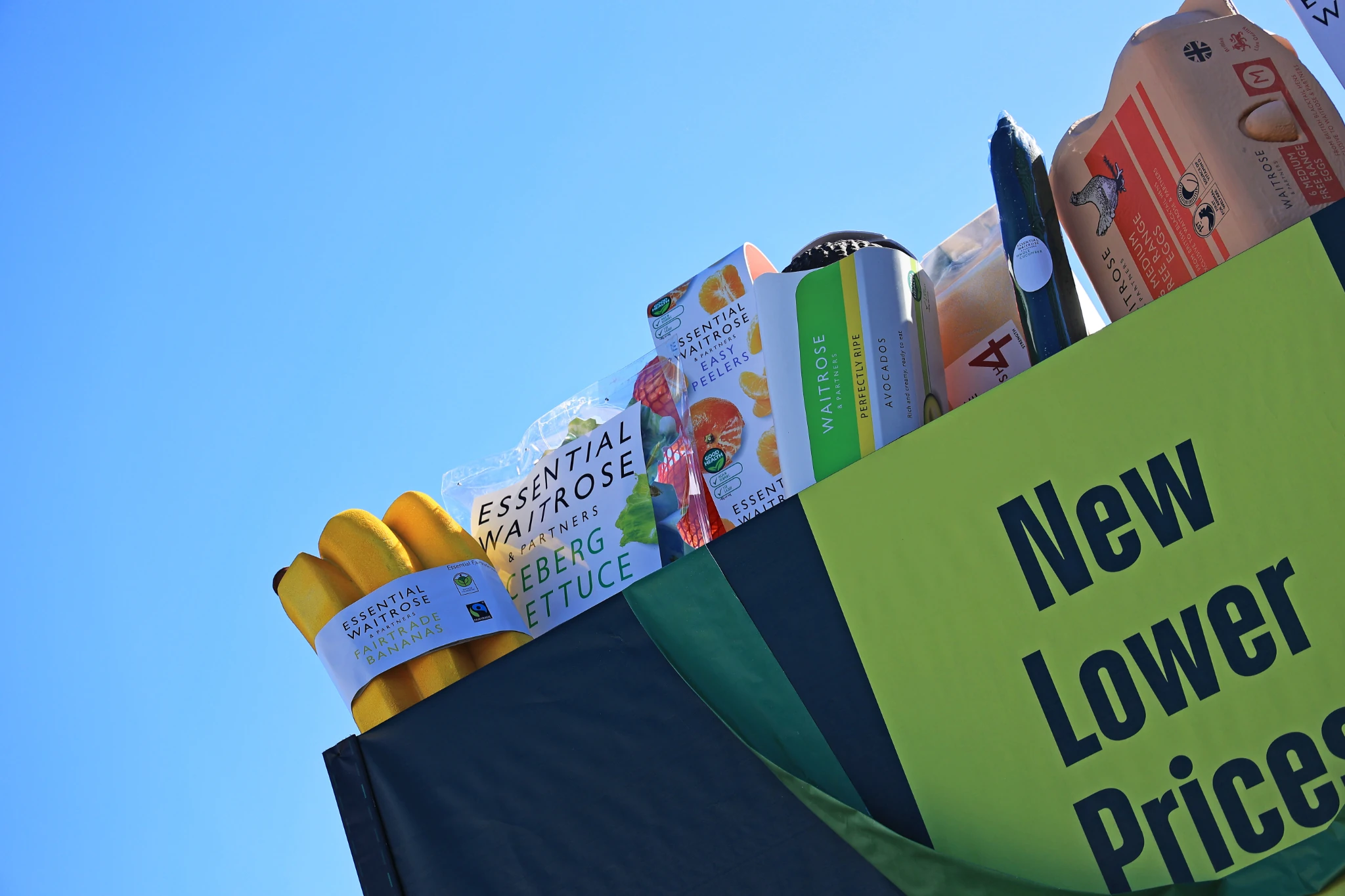 Close-up view of a Waitrose special-build billboard showing oversized grocery products and the New Lower Prices message against a blue sky.