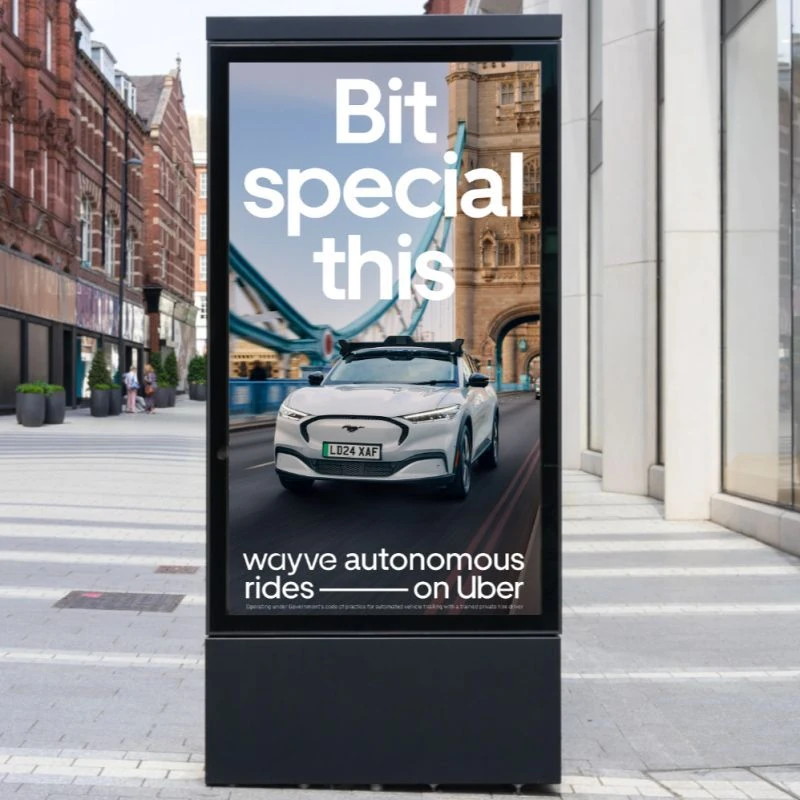 Street-level digital advertising panel in London showing a white Wayve autonomous vehicle on Uber with the headline Bit special this.