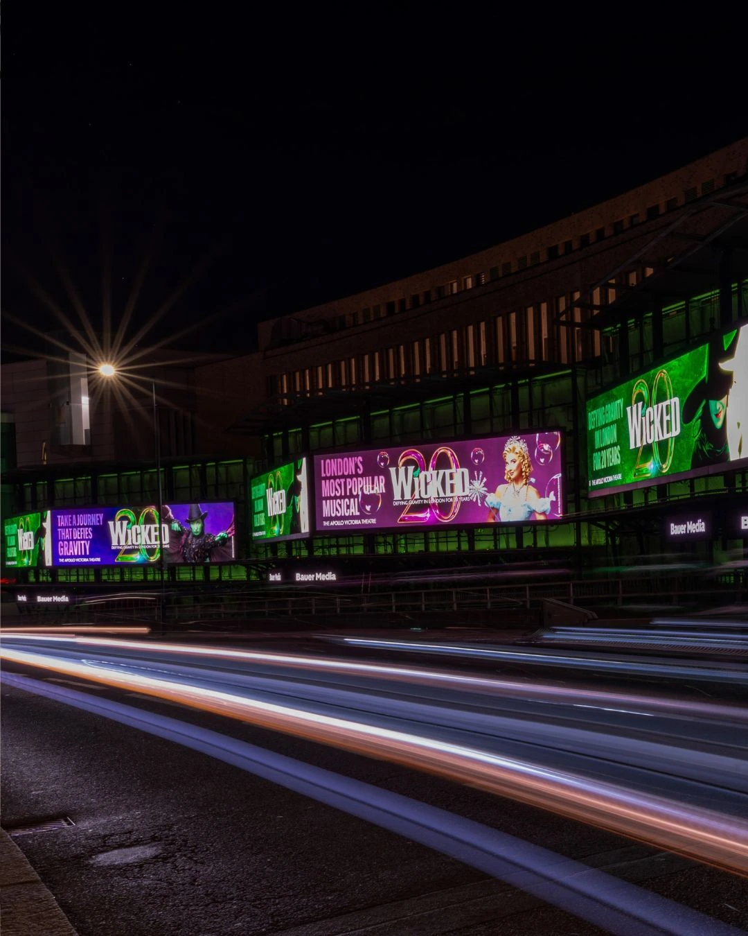 A row of large digital Wicked anniversary screens lighting up a London roadside at night.