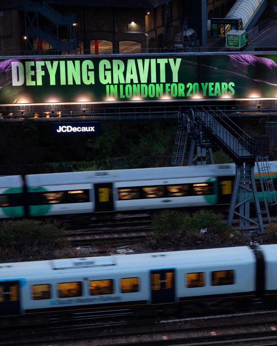 Large Wicked anniversary billboard in London reading Defying Gravity in London for 20 years above railway tracks near Victoria.