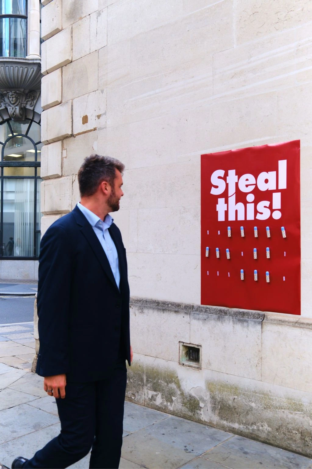 Businessman walking past a red Steal this poster featuring Wild refillable tinted lip balm samples attached to the wall.